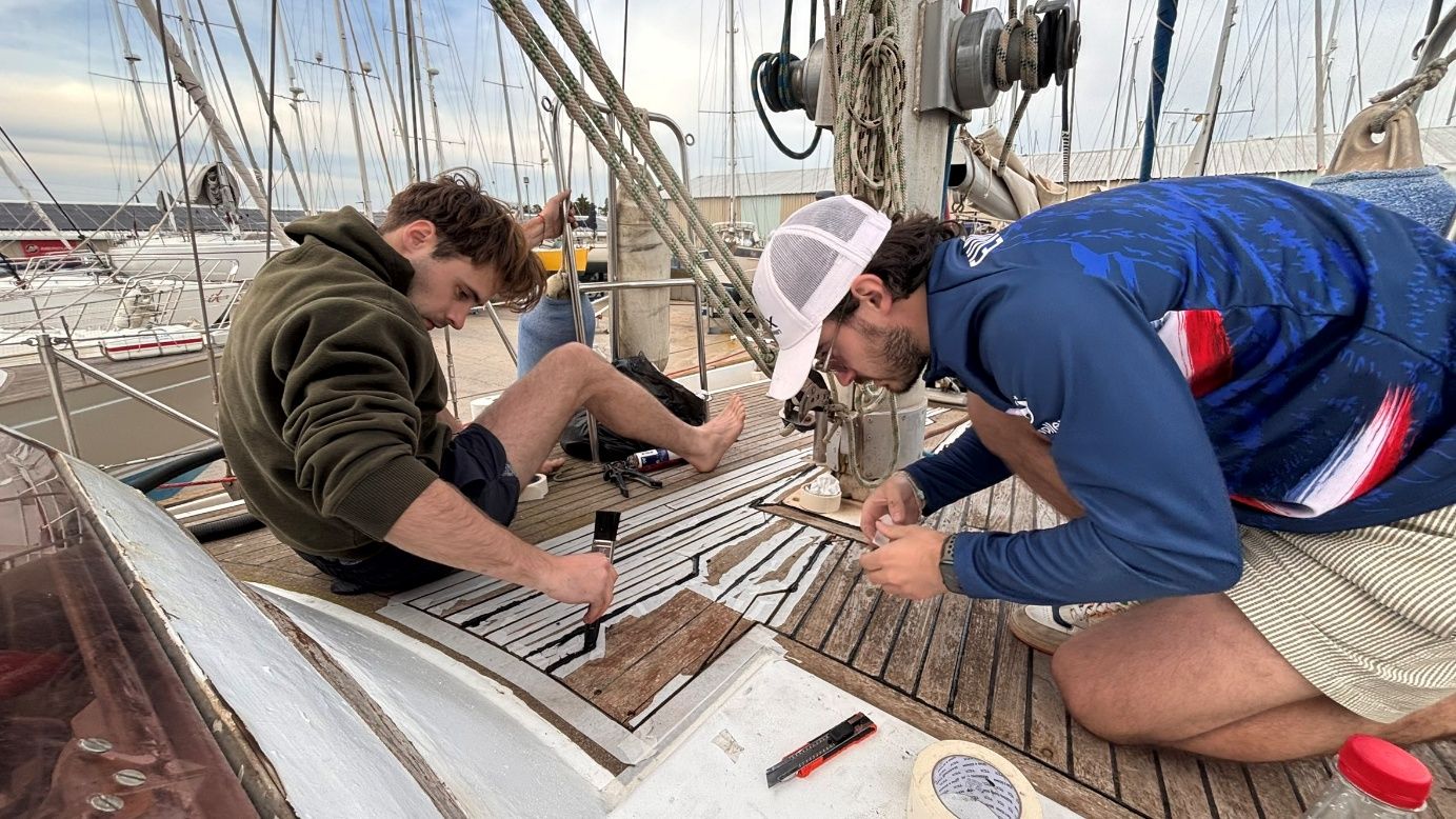 Arthur et Brieuc à genoux sur le pont, en train de refaire les joints Sika entre les lames de teck